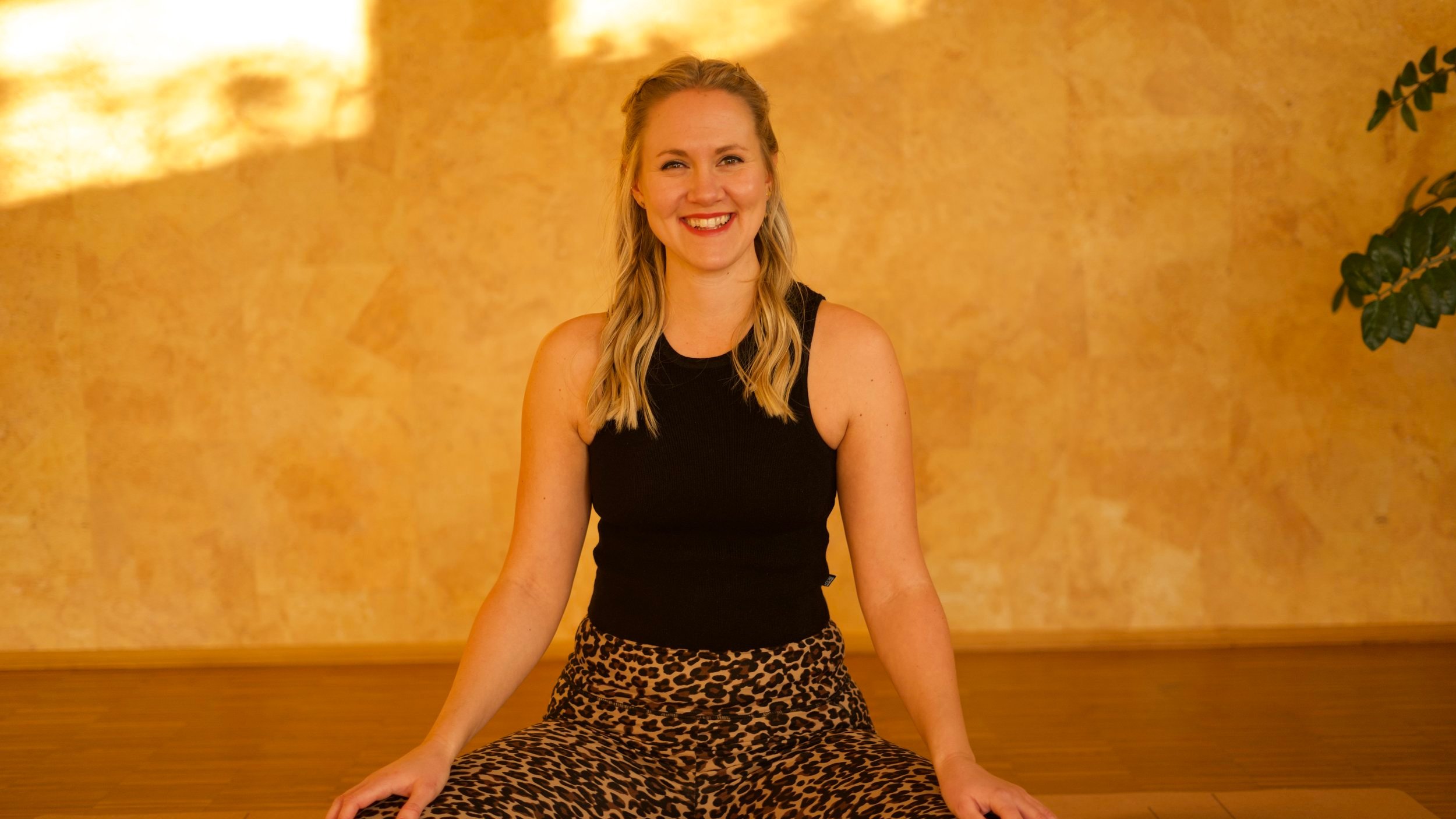 Lisa Becker sitzt lächelnd im Yogastudio vor einer goldgelben Wand